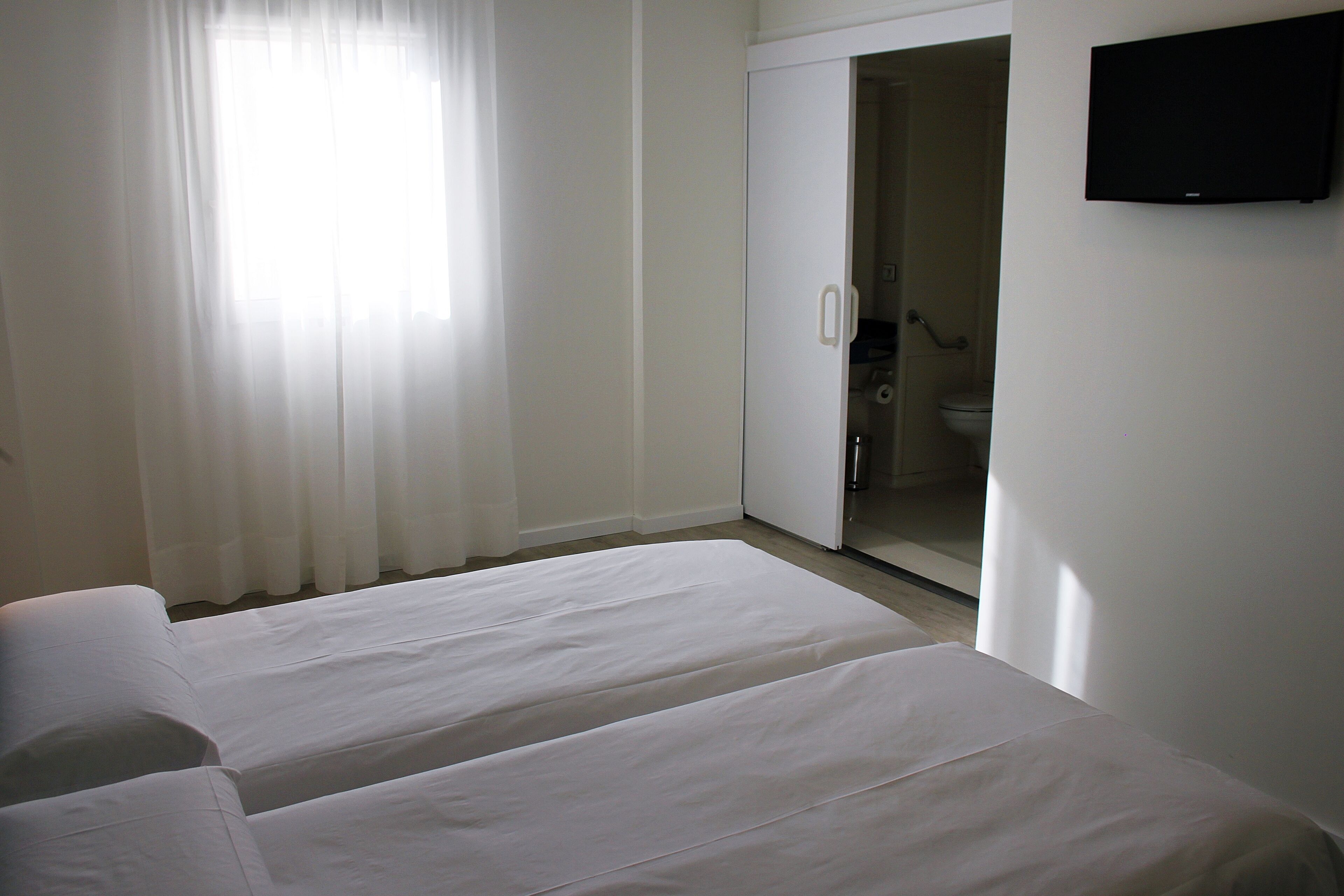 A hotel room with two beds, a bathroom with a toilet and shower, and a television on the wall.