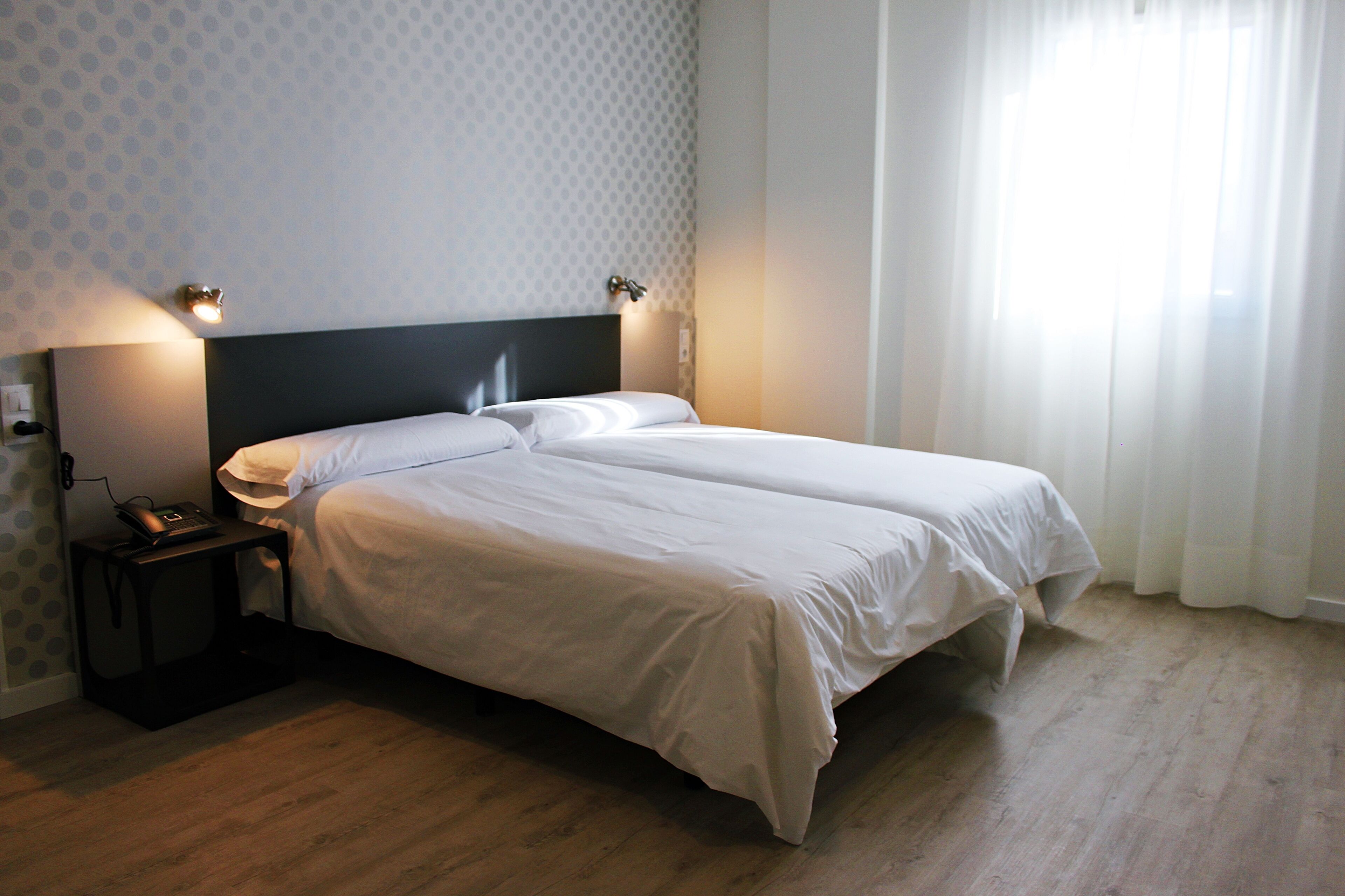 A neatly made double bed with white linens, a bedside table with a lamp, and a wall-mounted light fixture.