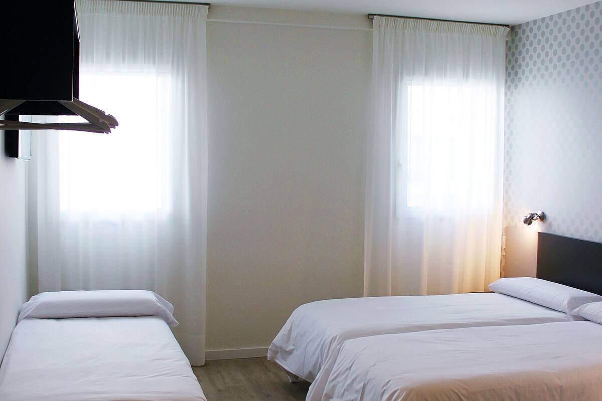 A hotel room with two single beds, a flat-screen TV mounted on the wall, and a window with sheer curtains.
