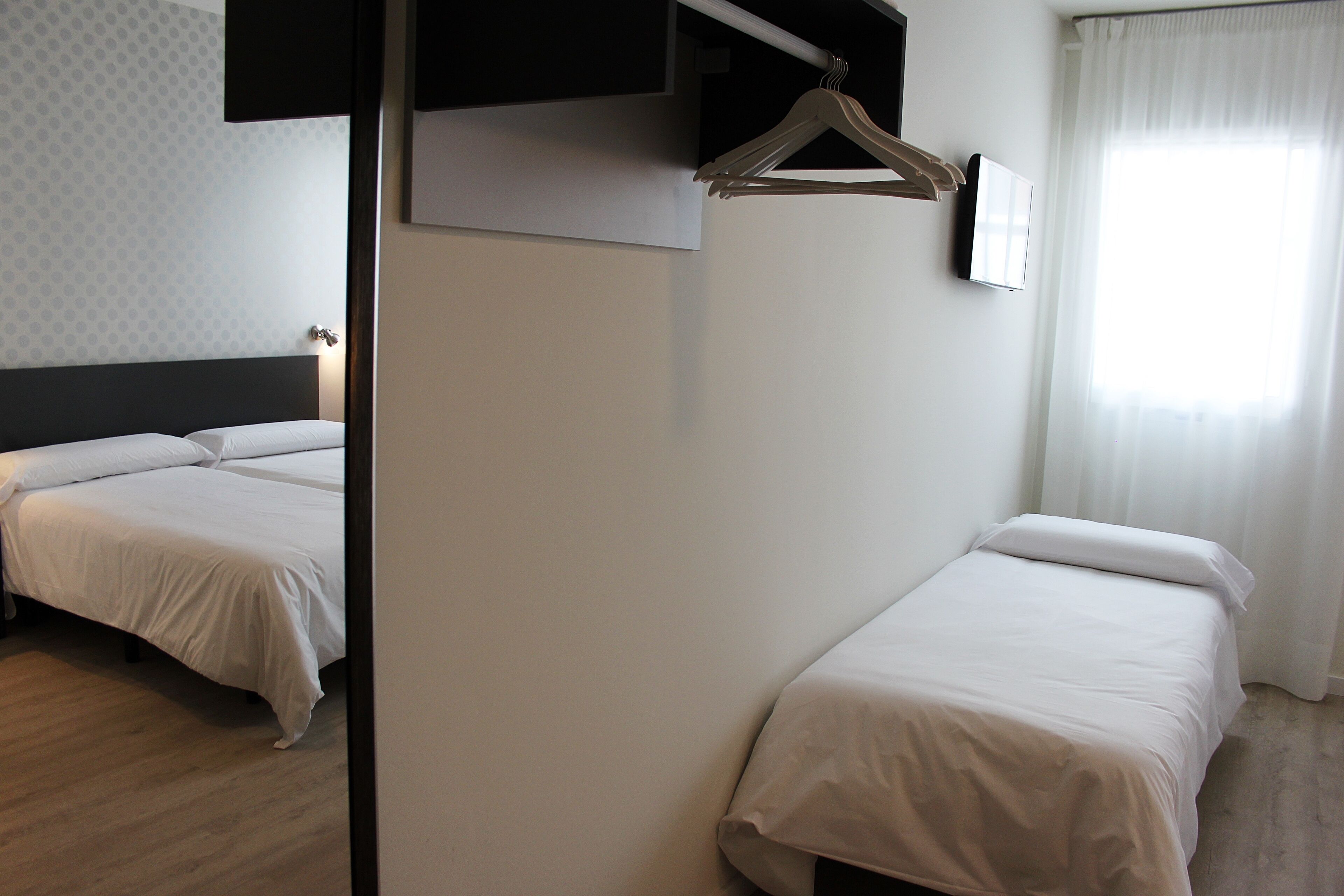 A hotel room with two beds, a wooden floor, a white wall, and a window with curtains.