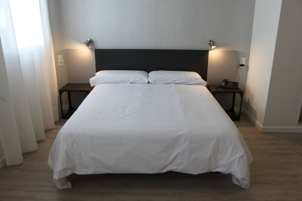 A neatly made bed with white linens in a room with a patterned wallpaper and a wooden floor.