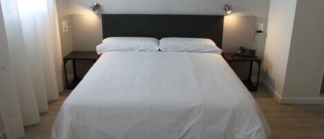 A large bed with a dark headboard, two bedside tables with lamps, and a plain wall with a geometric pattern.