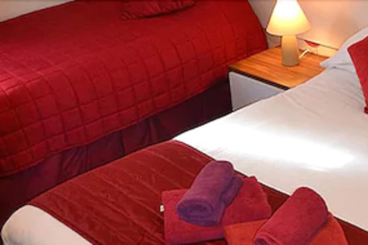 A hotel room with two beds, each with red bedding and pillows, a nightstand with a lamp, and a small table.