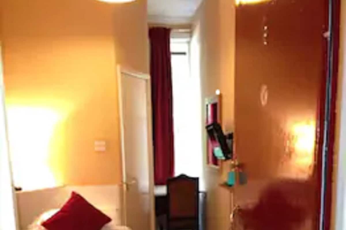 A narrow hotel corridor with a bed on the left, a door on the right, and a chair in the middle.