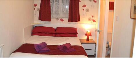 A bedroom with a bed, red curtains, a nightstand with a lamp, and a door to another room.