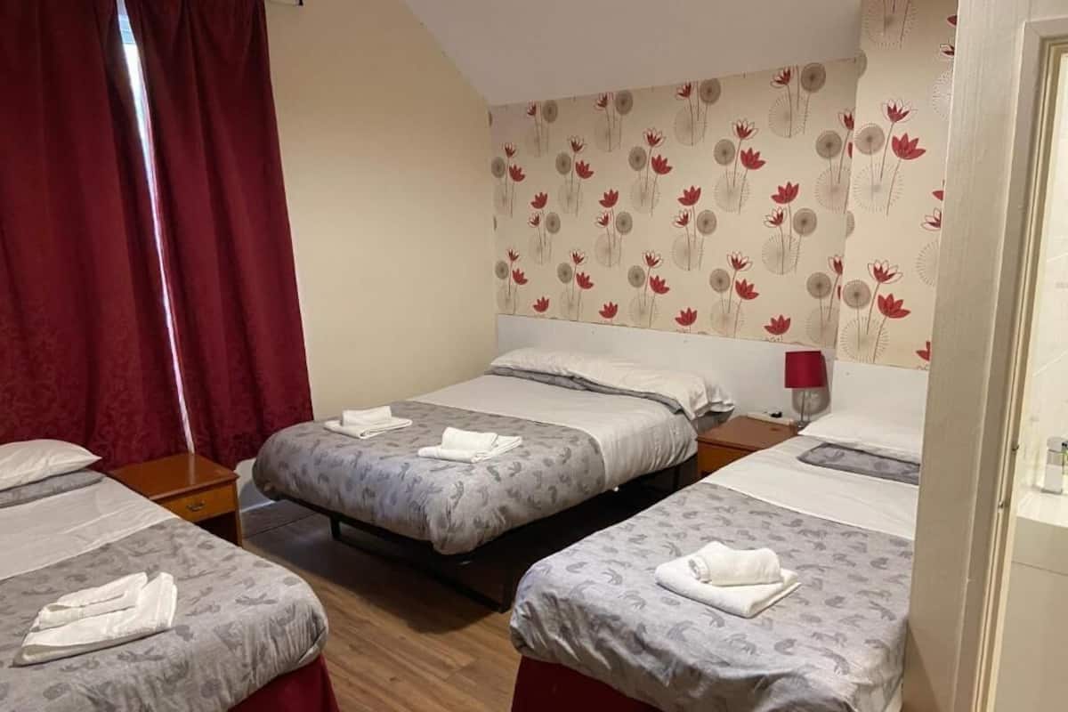 A hotel room with three beds, each with a grey floral bedspread and white pillows. There is a red curtain on the left side, and a wall with a floral wallpaper pattern.