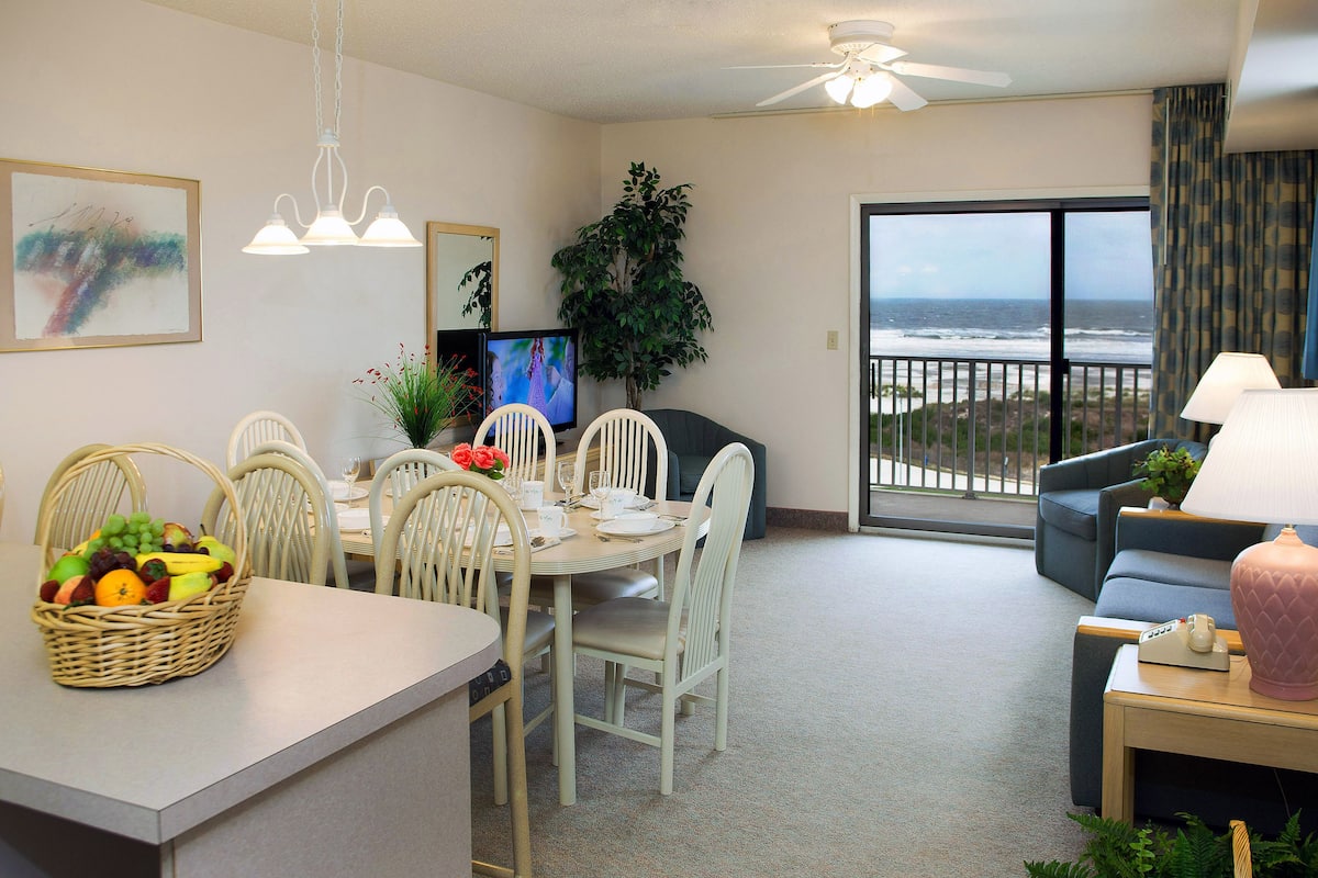 A dining area with a table set for four, a television, and a large window with a view of the ocean.