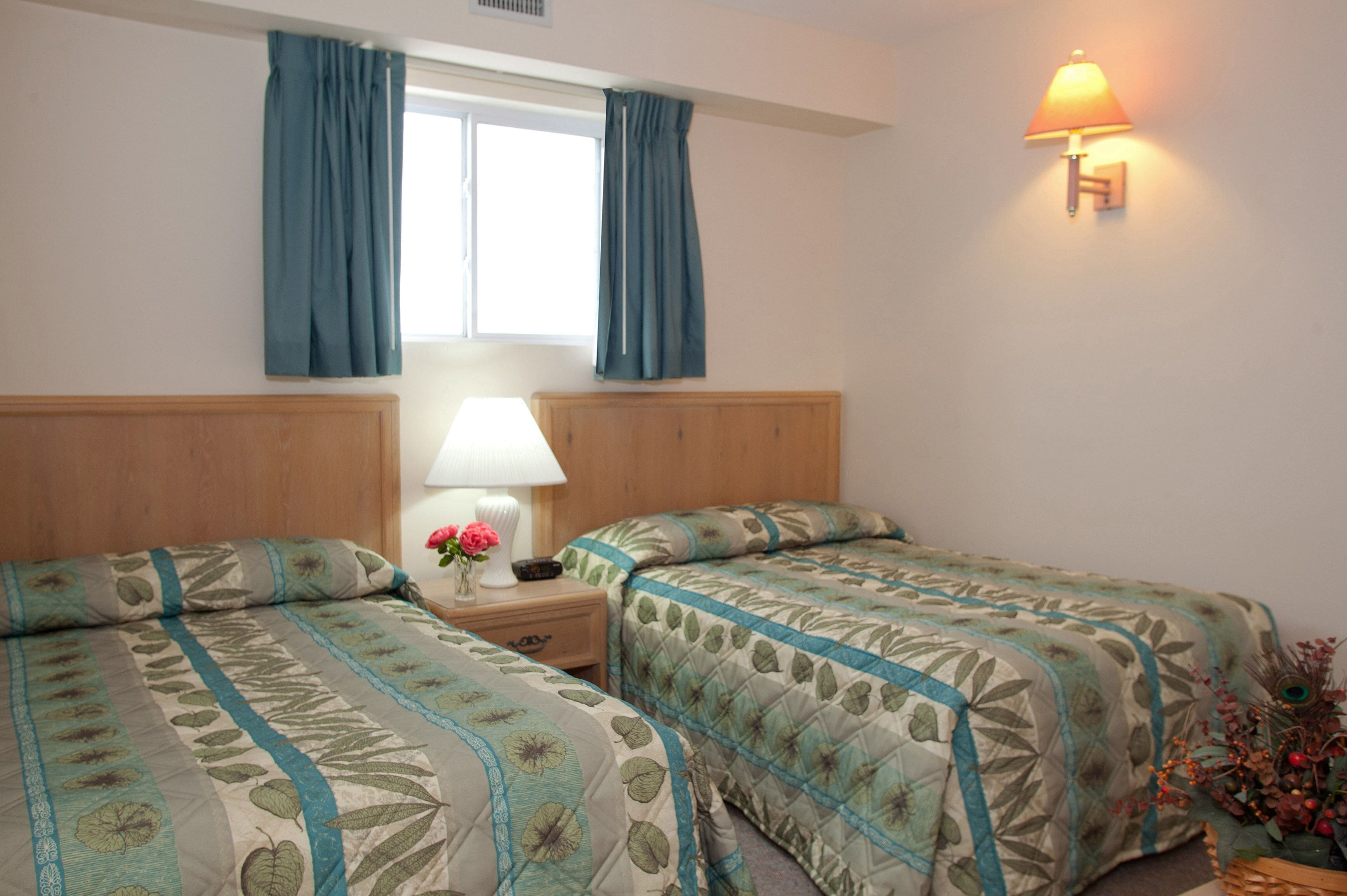 A hotel room with two beds, a nightstand with a lamp, a window with curtains, and a wall-mounted light fixture.