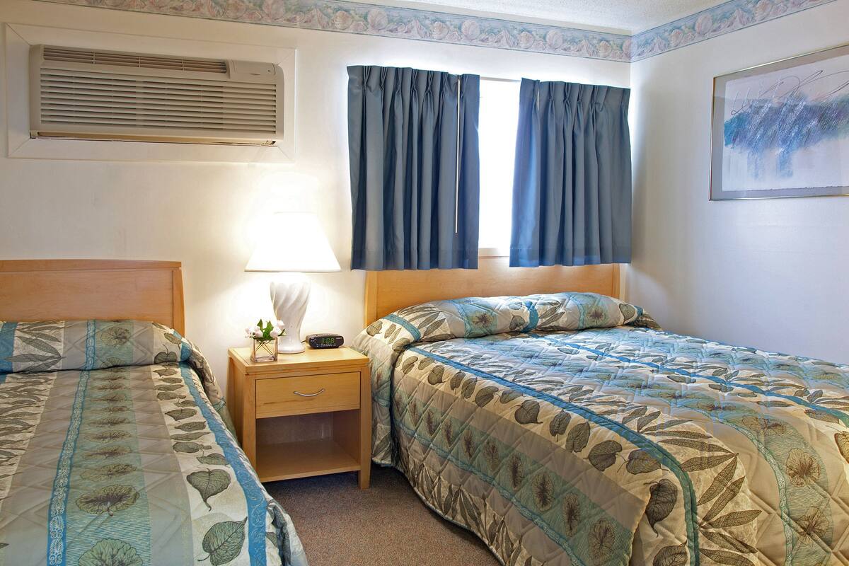 A hotel room with two beds, a nightstand with a lamp, a window with curtains, and a wall-mounted air conditioner.