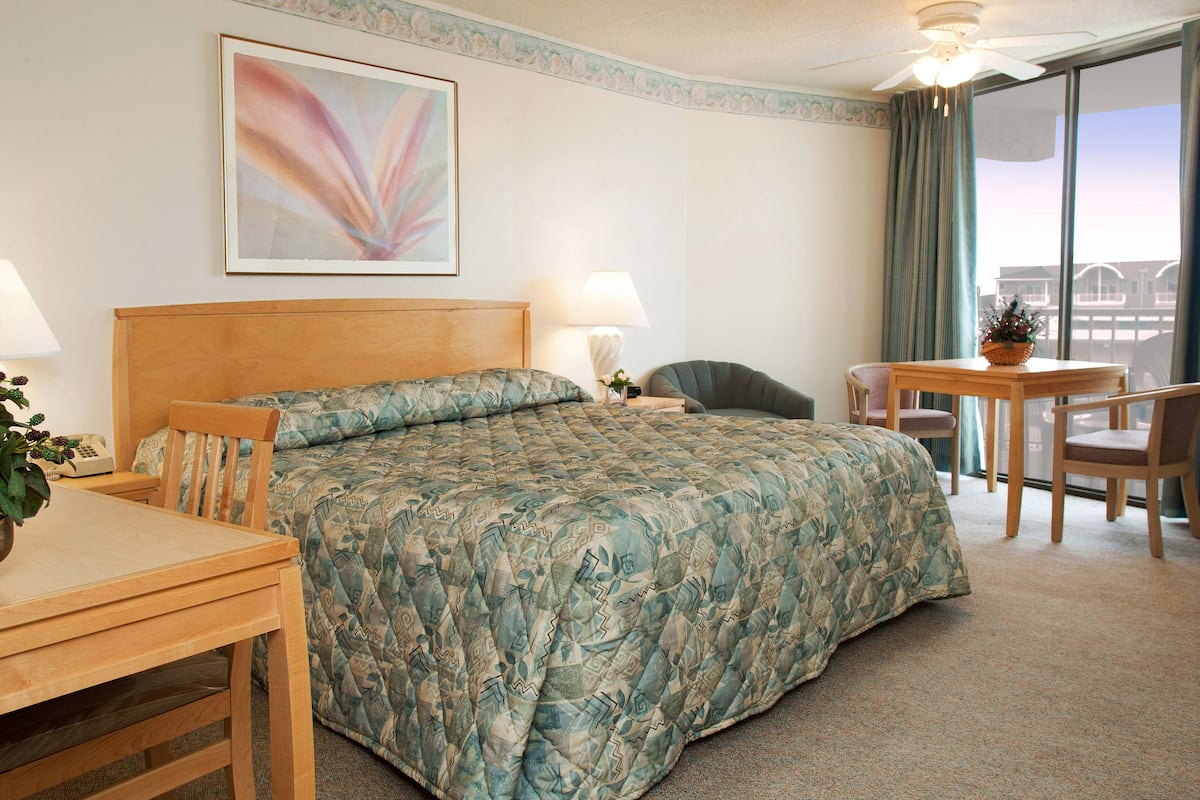 A hotel room with a bed, a desk, a chair, a table, a lamp, a ceiling fan, a window with curtains, and a framed picture on the wall.