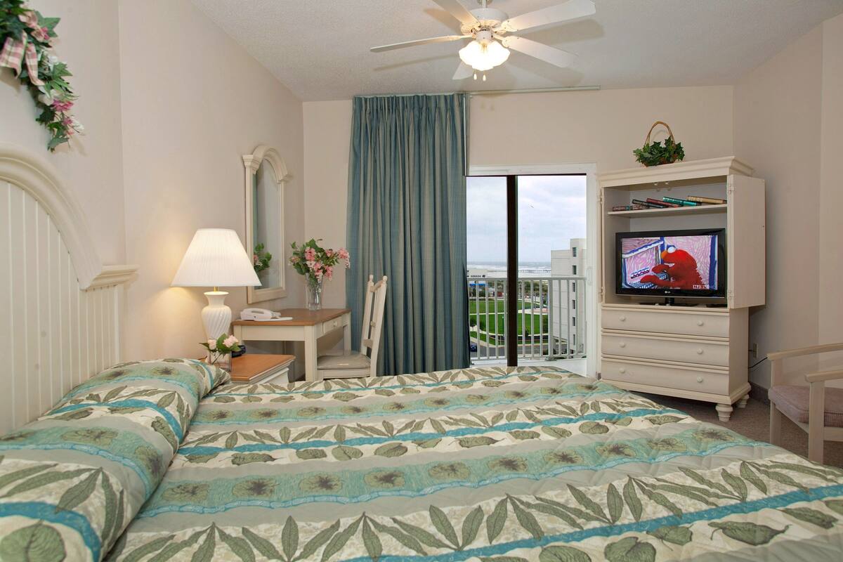 A hotel room with a bed, a television, a desk, a chair, a window with a view, and a ceiling fan.