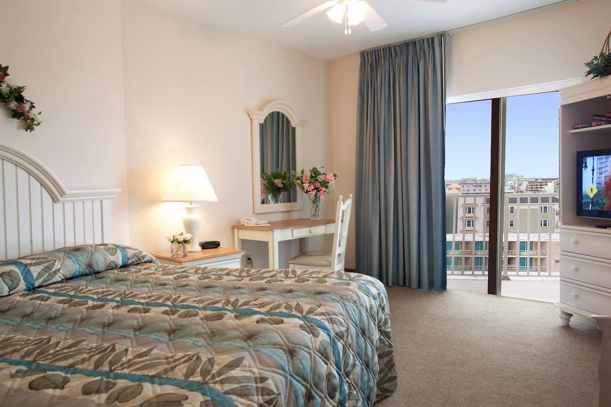 A hotel room with a bed, a desk with a lamp, a chair, a TV, and a balcony with a city view.