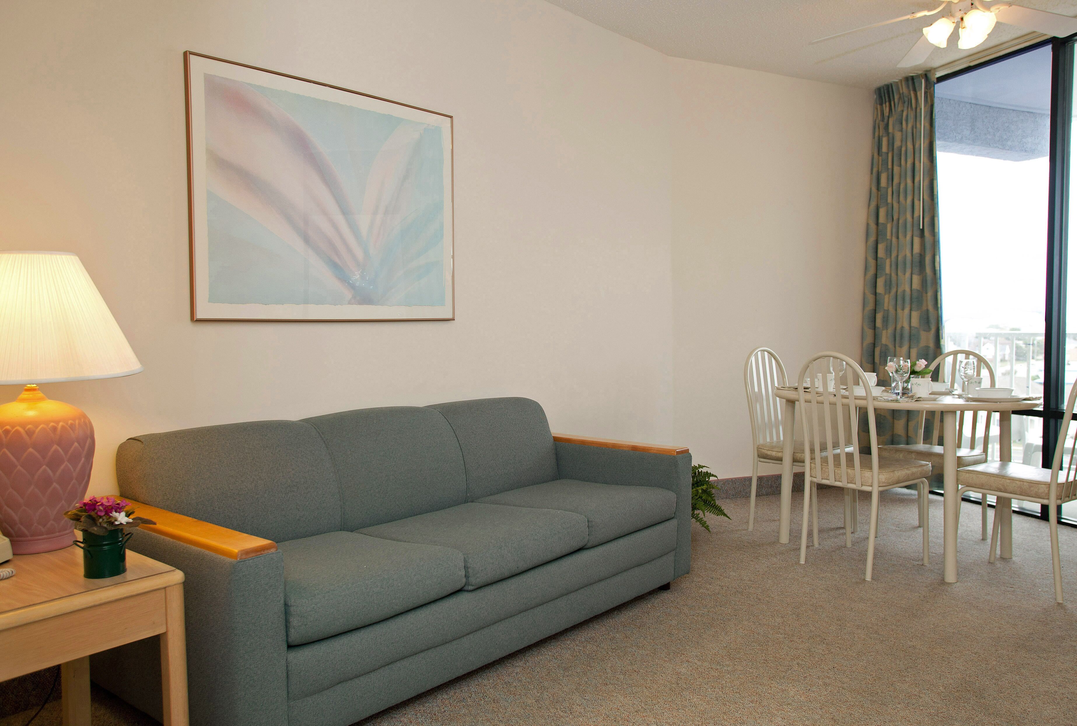 A modern living room with a grey sofa, a dining table with chairs, a lamp, and a window with curtains.