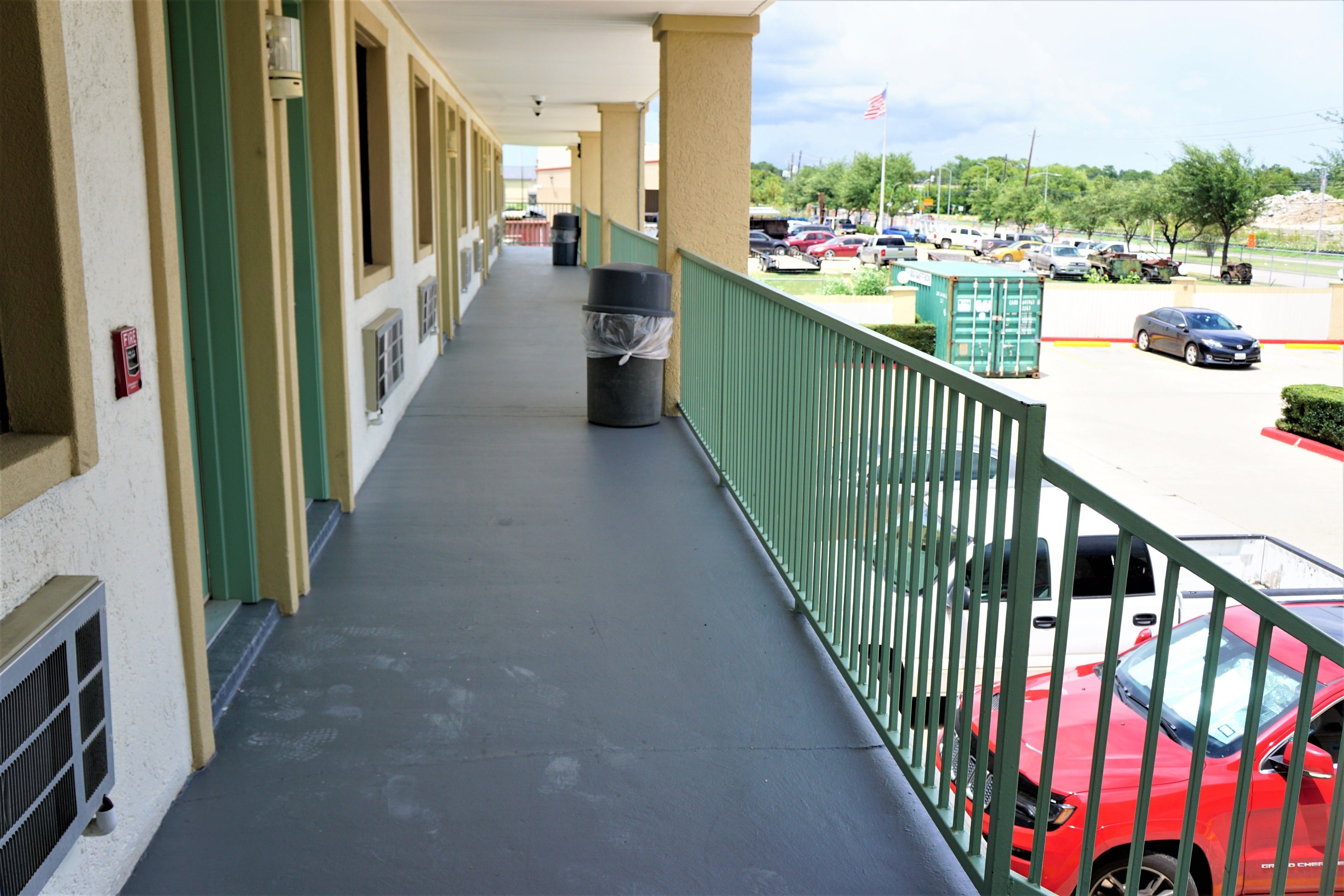 A hotel corridor with a covered walkway, a trash bin, and a view of a parking lot with various cars.