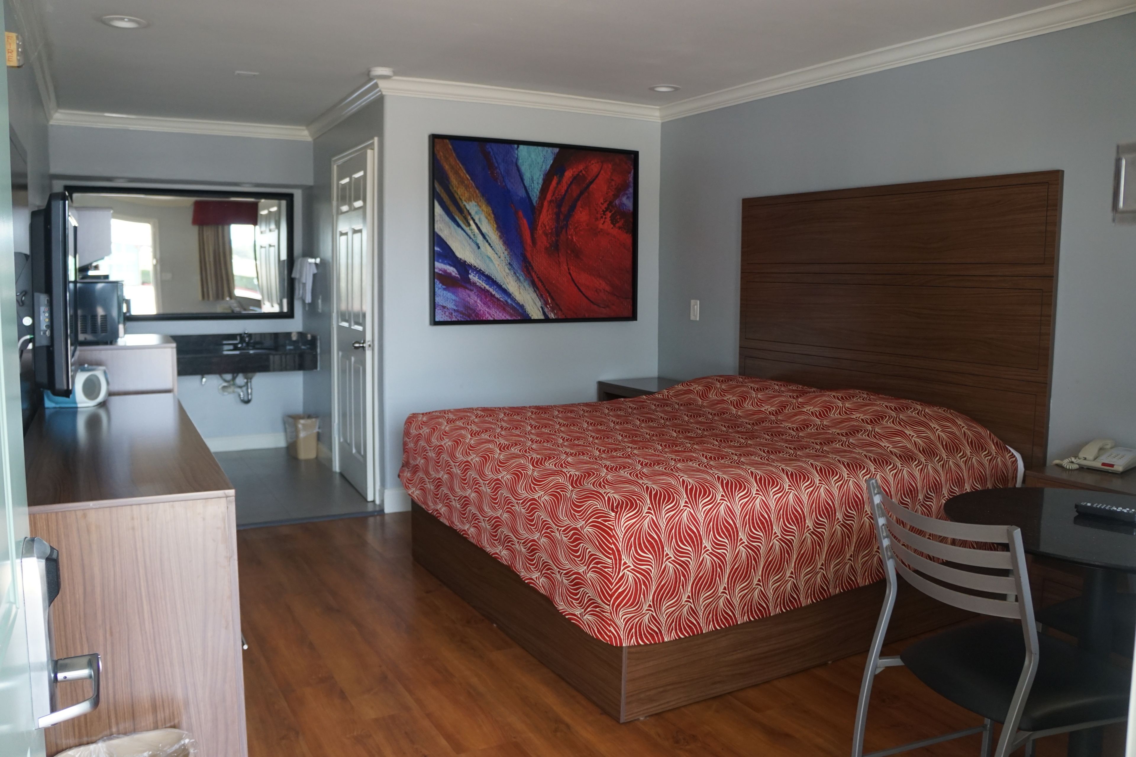 Habitación de hotel con cama de madera, un escritorio, una silla, un cuadro abstracto grande y un baño visible a través de una puerta abierta.