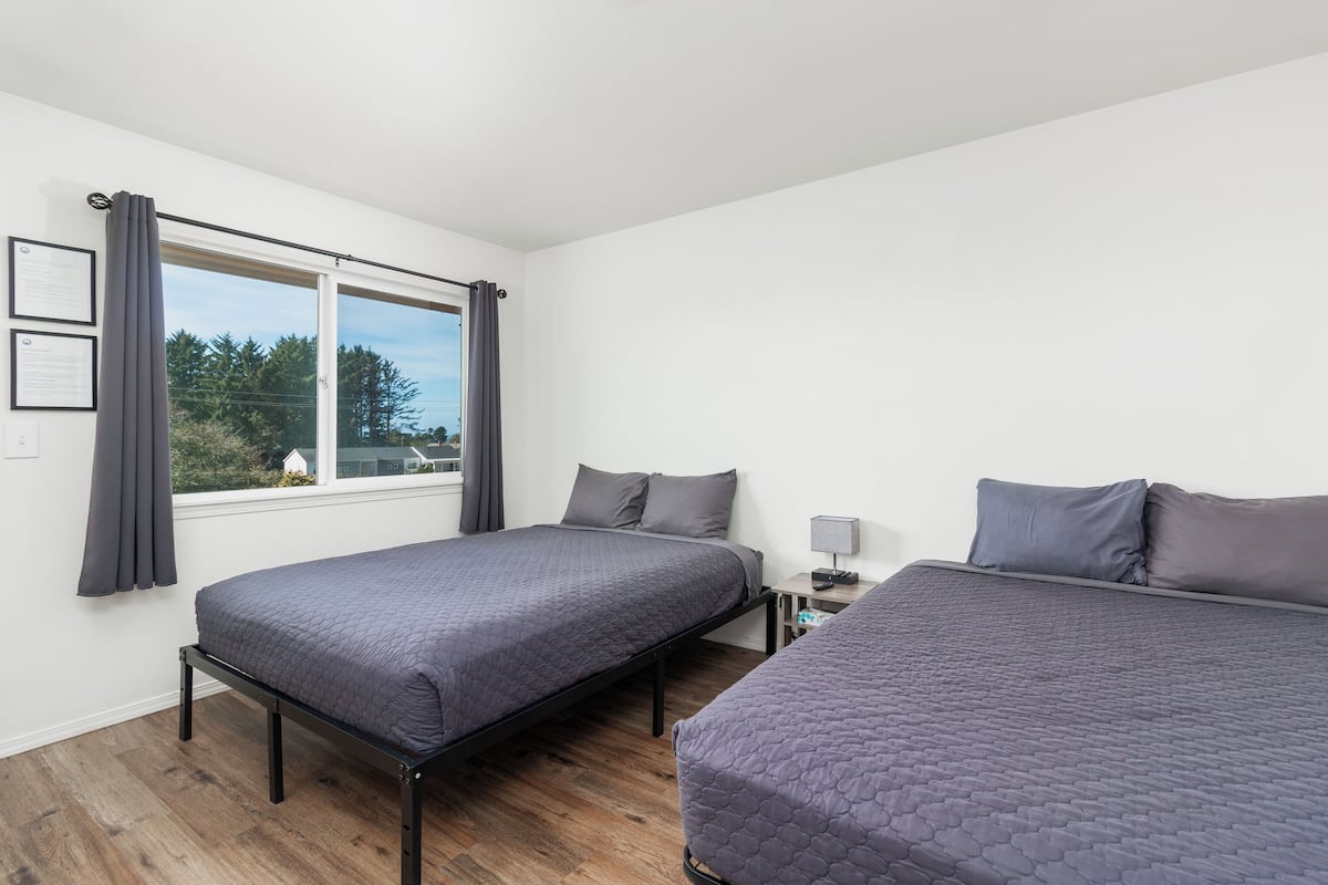 A room with two beds, a window with curtains, a nightstand with a lamp, and a view of trees outside.