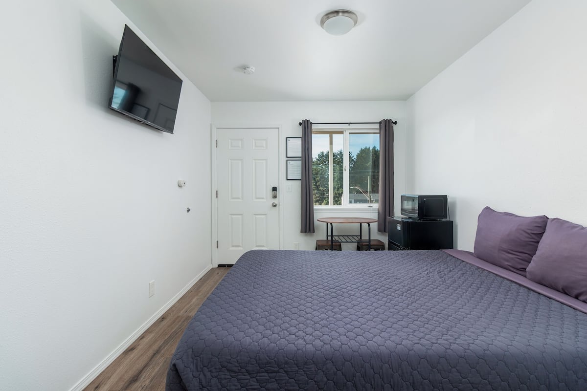 A bedroom with a bed, a TV mounted on the wall, a small table with a chair, and a window with curtains.