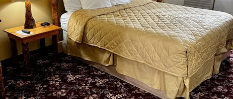 Une chambre d’hôtel avec un grand lit, une tête de lit en bois, un tableau encadré au mur, une table de chevet avec une lampe et une moquette à motifs floraux.