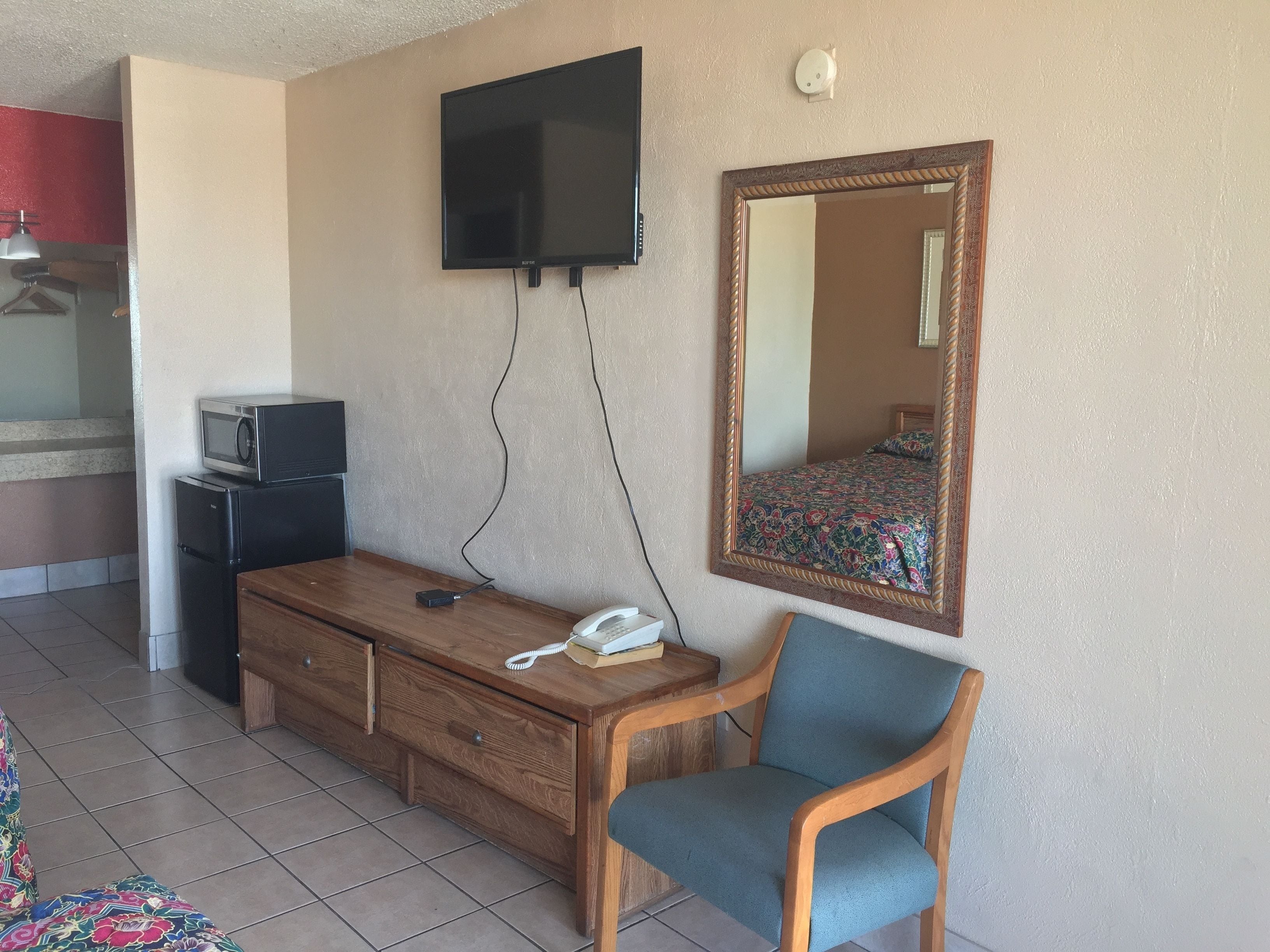 A hotel room with a flat-screen TV mounted on the wall, a wooden dresser, a blue chair, a small desk with a telephone, and a microwave on a cabinet.