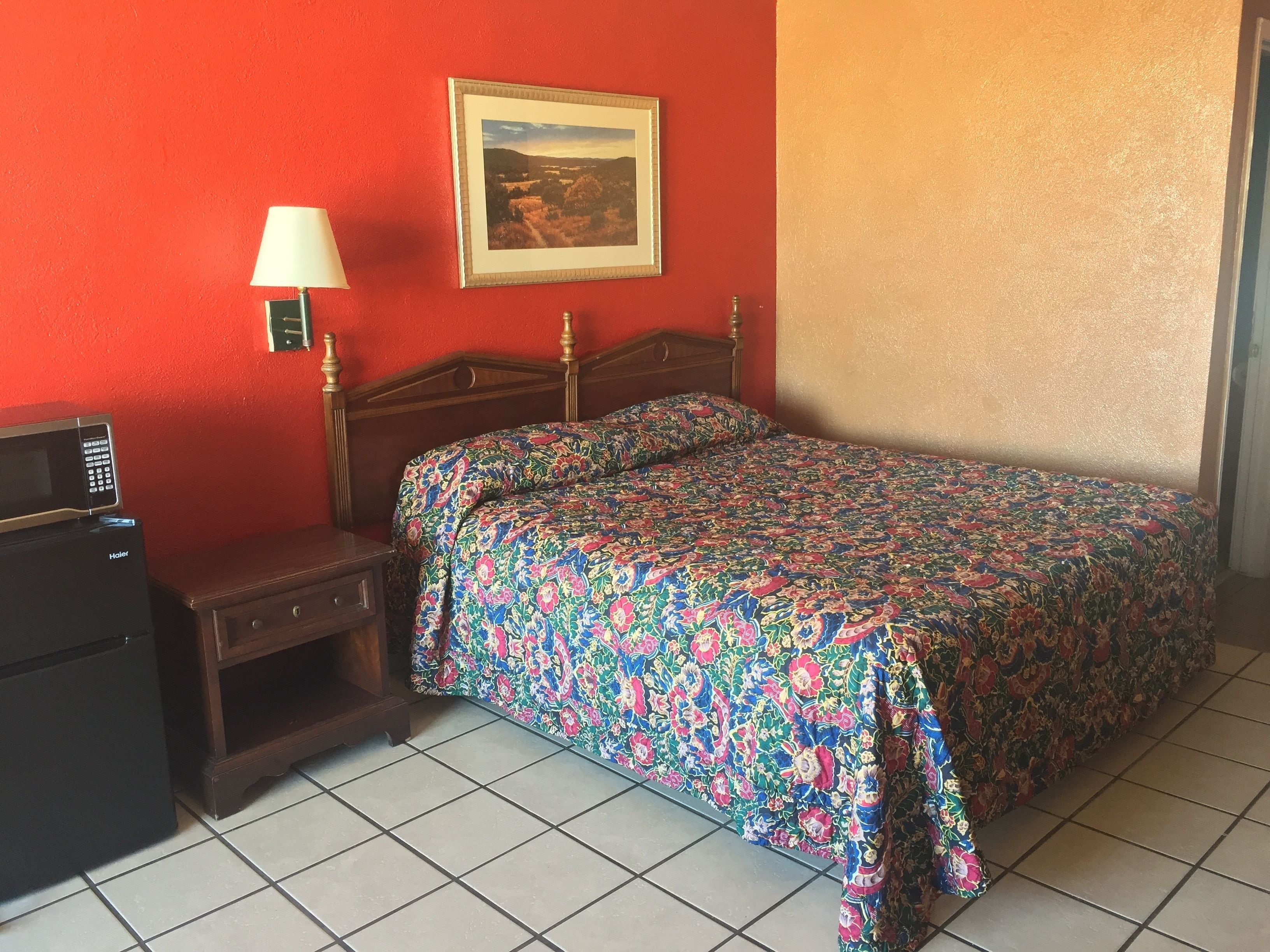 A bedroom with a bed, a nightstand, a lamp, a framed picture on the wall, and a microwave on the dresser.