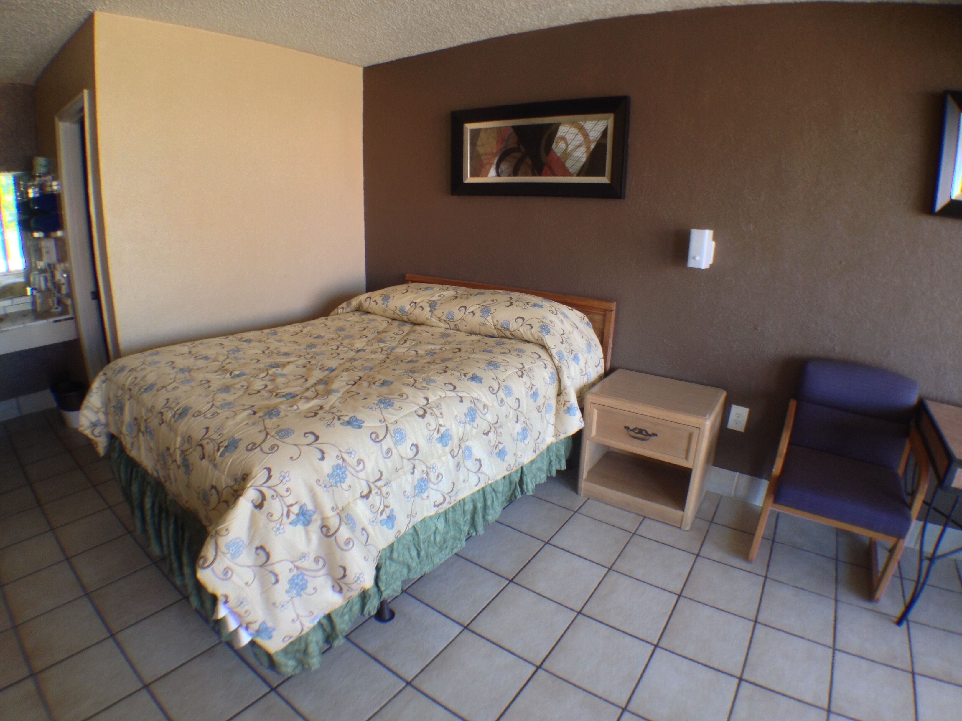 Un dormitorio con cama, mesita de noche, una silla y un cuadro en la pared.