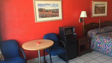 A hotel room with a red wall, a bed with a floral bedspread, a small table with a red cup, a black heater, and two framed pictures on the wall.