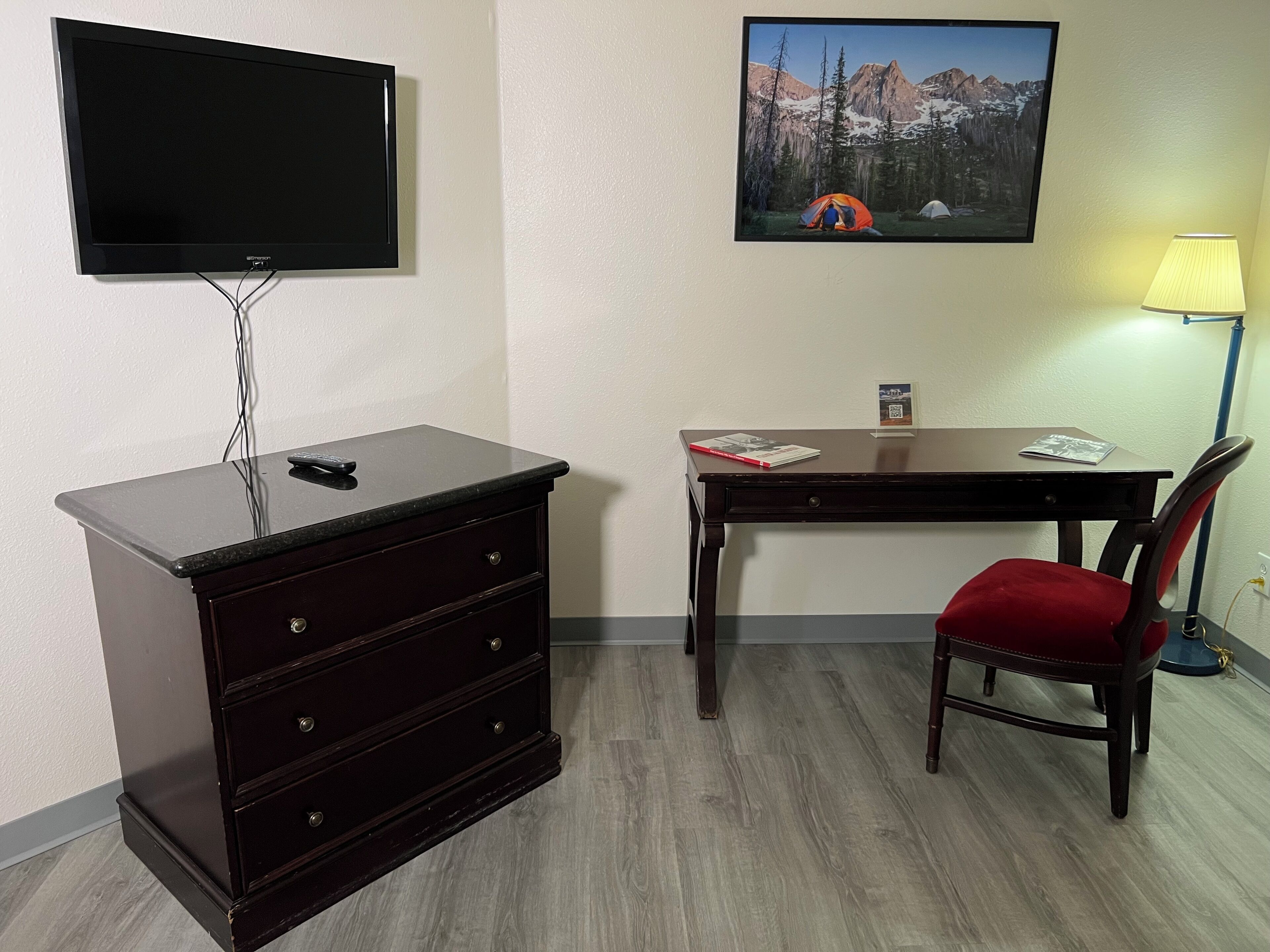 A room with a TV mounted on the wall, a desk with a chair, a dresser, and a lamp.