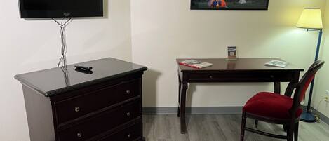 A room with a TV mounted on the wall, a desk with a chair, a dresser, and a lamp.