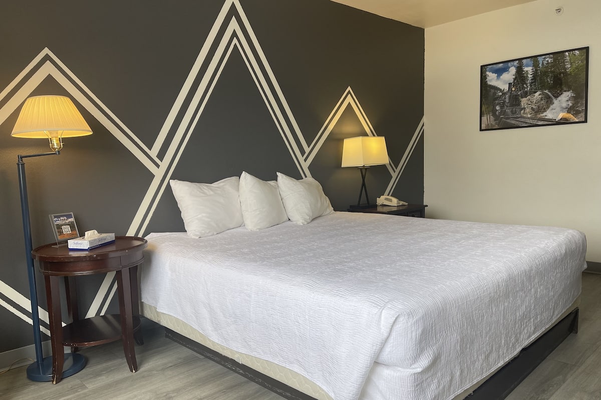 Une chambre d’hôtel avec un grand lit, deux lampes de chevet, une table de chevet avec un téléphone, un tableau encadré au mur et une petite table avec une lampe et une brochure.