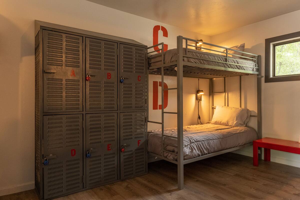 Un lit superposé avec une armoire en métal, un banc rouge et une fenêtre laissant entrer la lumière naturelle.