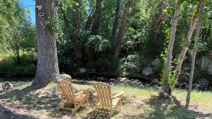 Deux fauteuils Adirondack en bois, placés dans un cadre naturel, avec un tronc d’arbre et un ruisseau en arrière-plan.