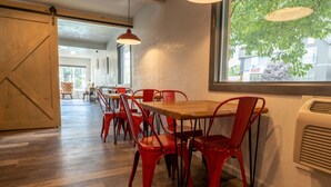 Un espace repas avec des tables en bois et des chaises rouges, une grande fenêtre donnant sur les arbres et une porte coulissante de style grange.