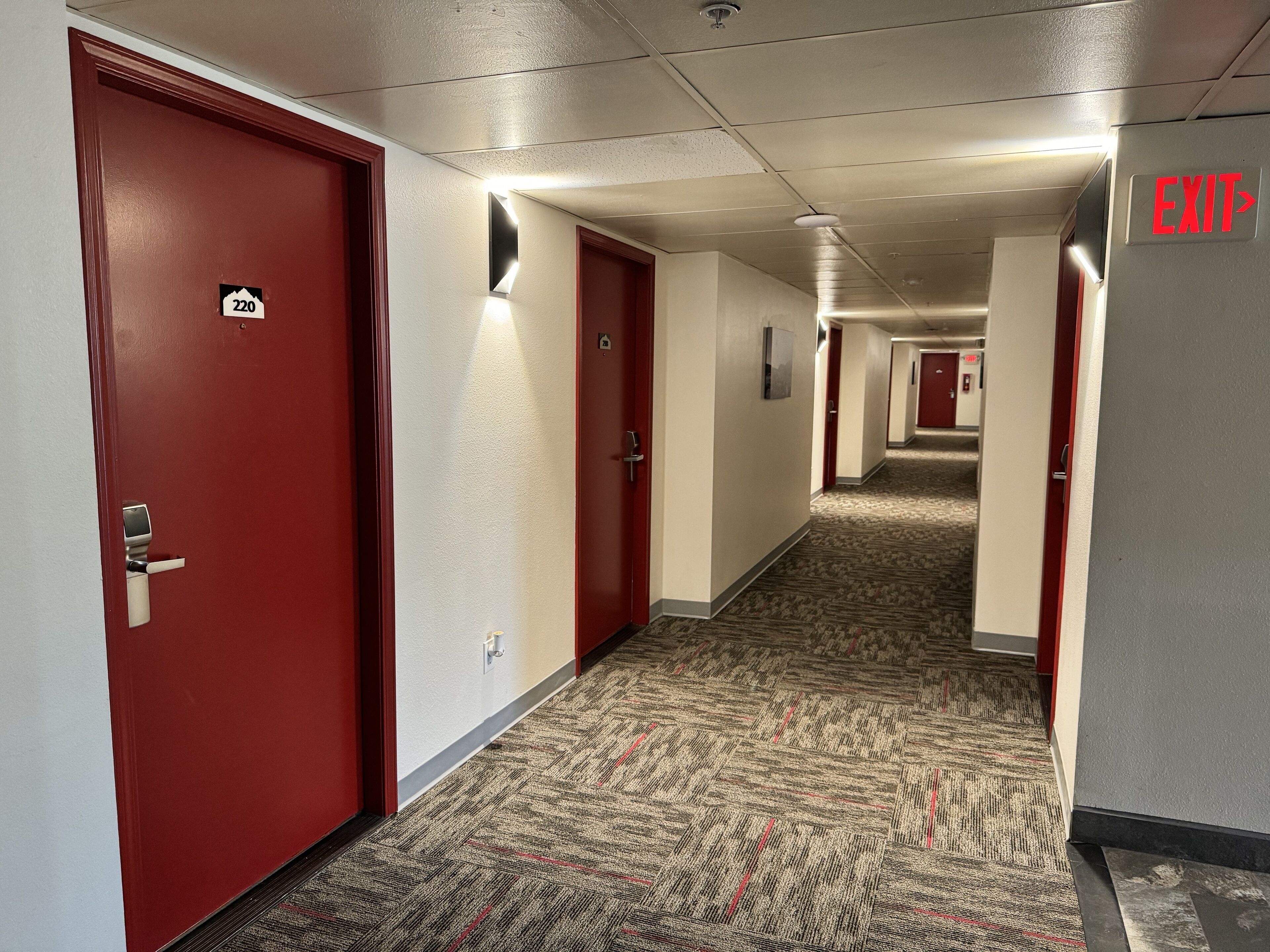 A hotel corridor with red doors, carpeted floors, and an EXIT sign.