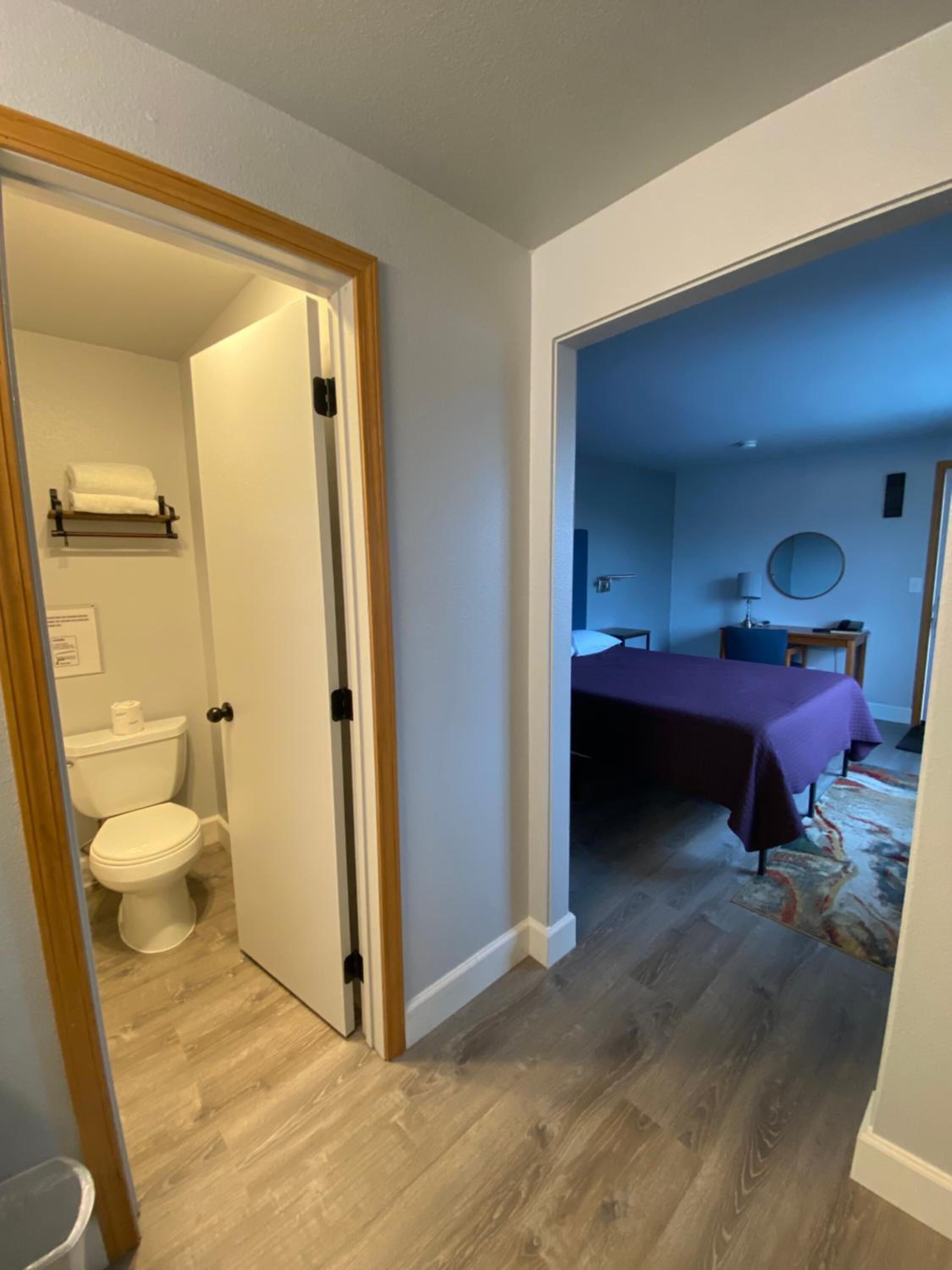 Una habitación con piso de madera, una cama con colcha morada, una mesita y un espejo en la pared.