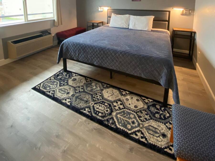 A bedroom with a bed, a patterned rug, a nightstand, a wall-mounted artwork, and a window with curtains.