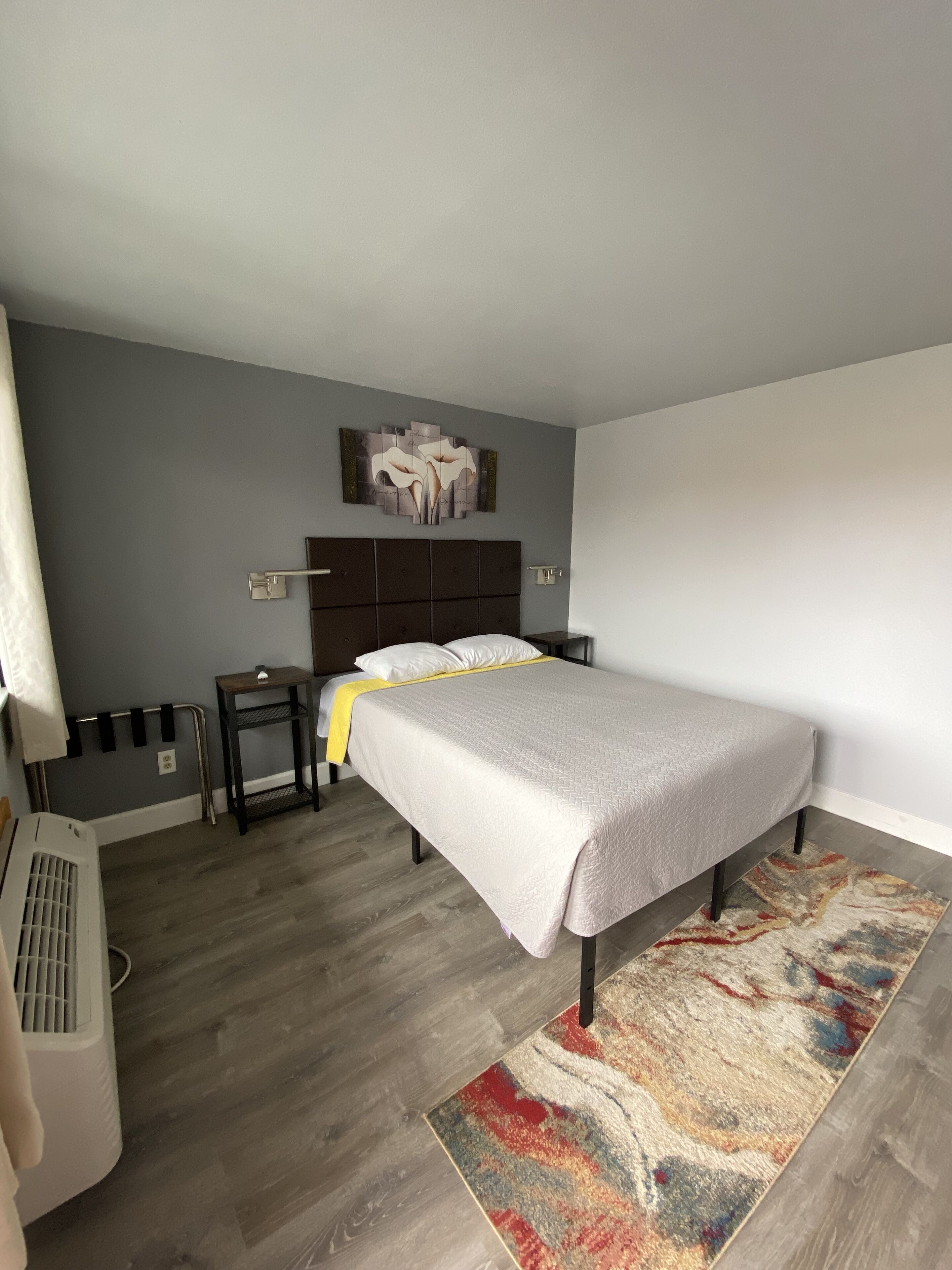 Een slaapkamer met een bed, nachtkastje, radiator en een kunstwerk aan de muur.