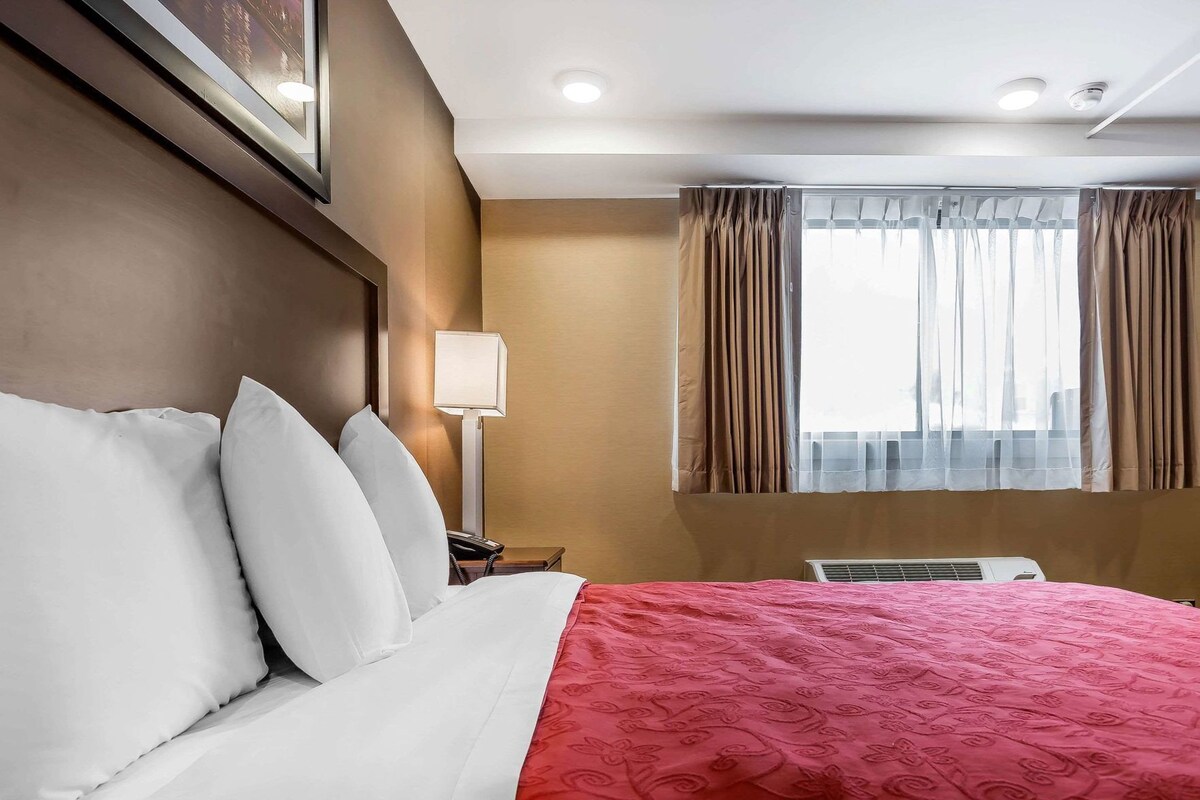 A hotel room with a bed, a bedside lamp, a telephone, a window with curtains, and an air conditioning unit.