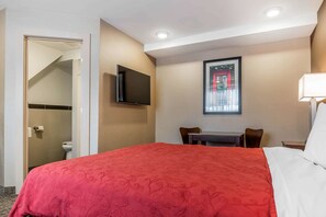A hotel room with a large bed, a television mounted on the wall, a desk with a chair, and a window with a view of a building.