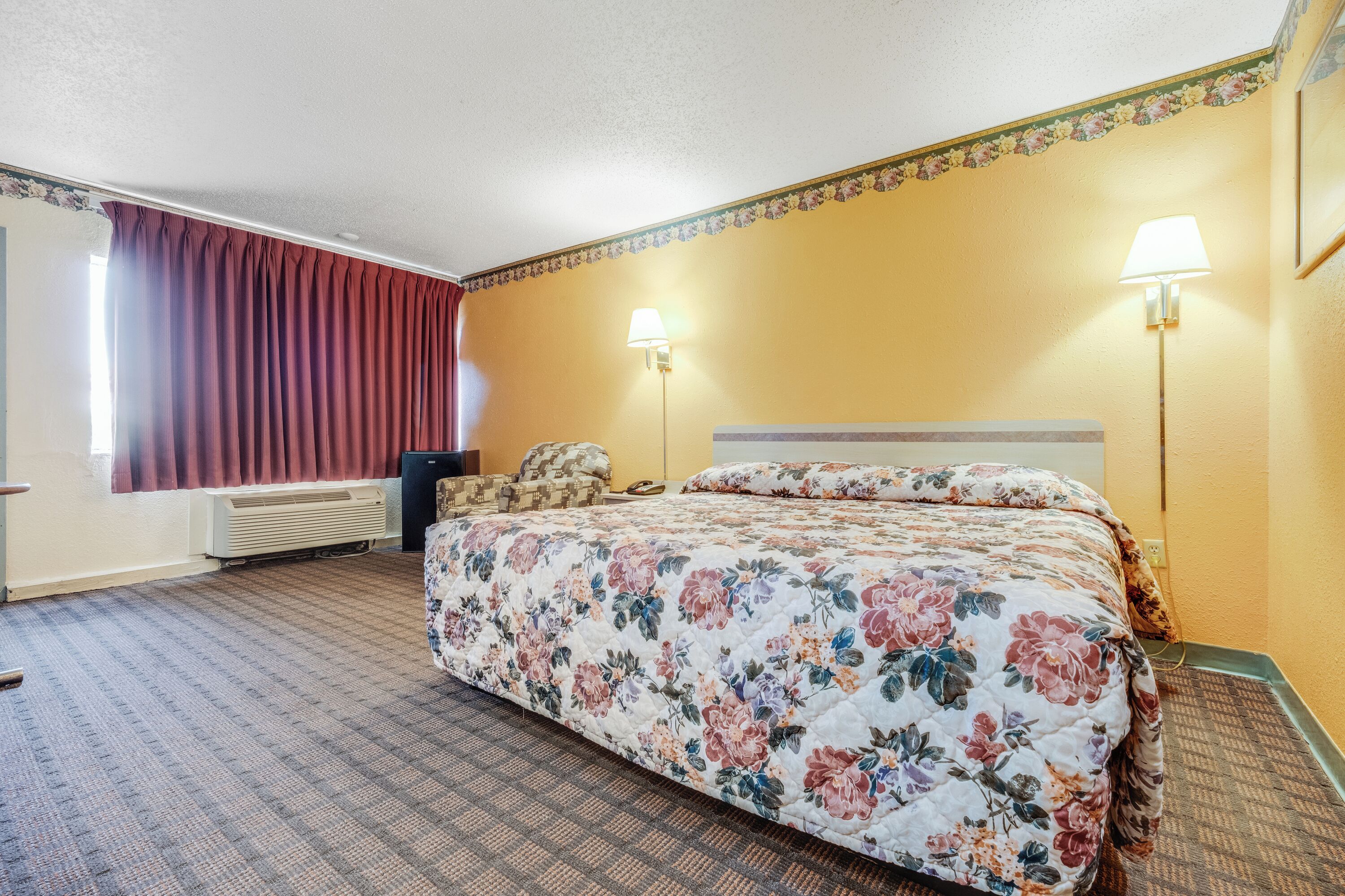 Une chambre d’hôtel avec une couvre-lit à motifs floraux, deux lampes de chevet, un radiateur et une fenêtre avec des rideaux rouges.