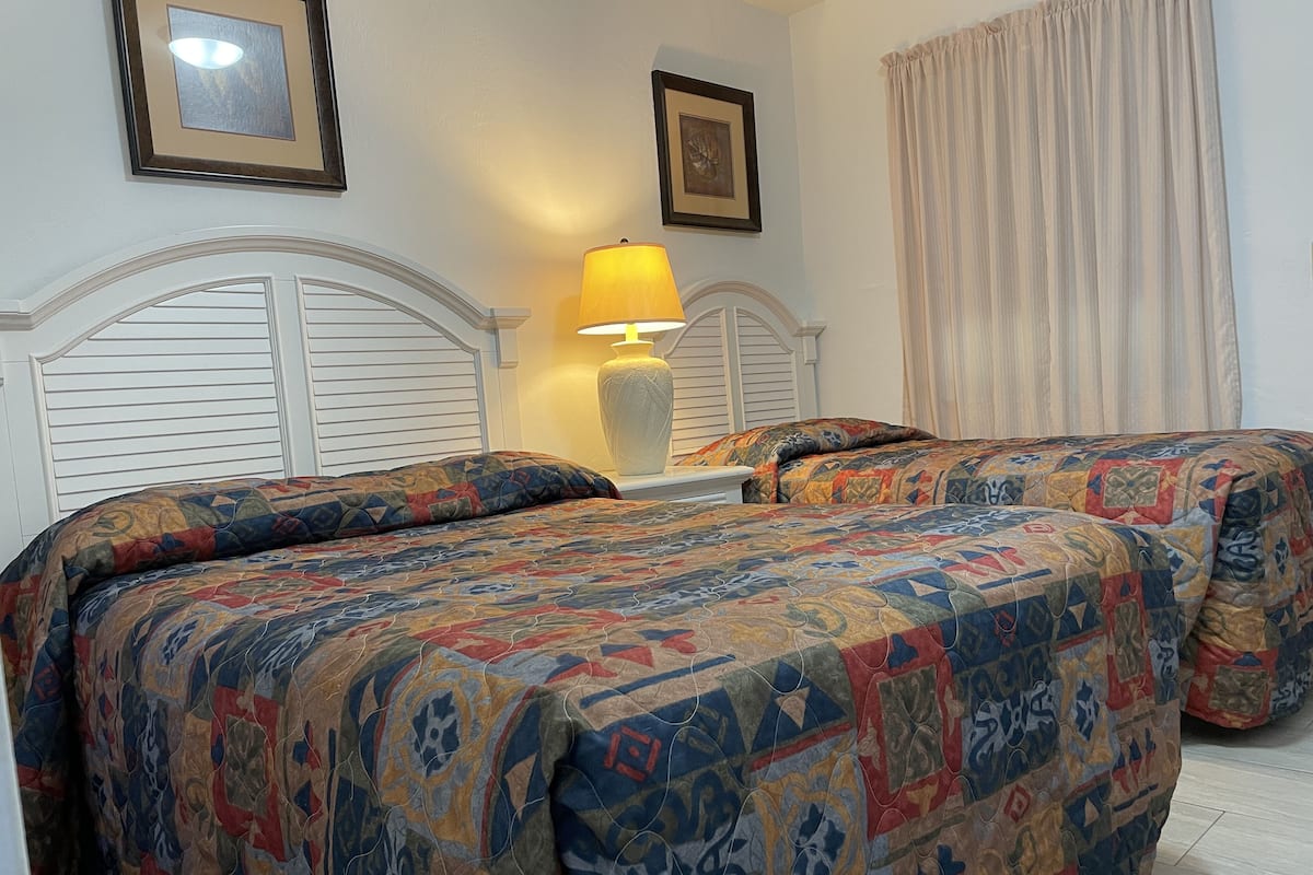 A bedroom with two beds, each with a patterned bedspread, a bedside table with a lamp, and framed artwork on the walls.
