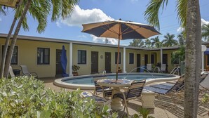 Un área de piscina con sillones de descanso y una sombrilla, rodeada de palmeras y un edificio con múltiples ventanas.