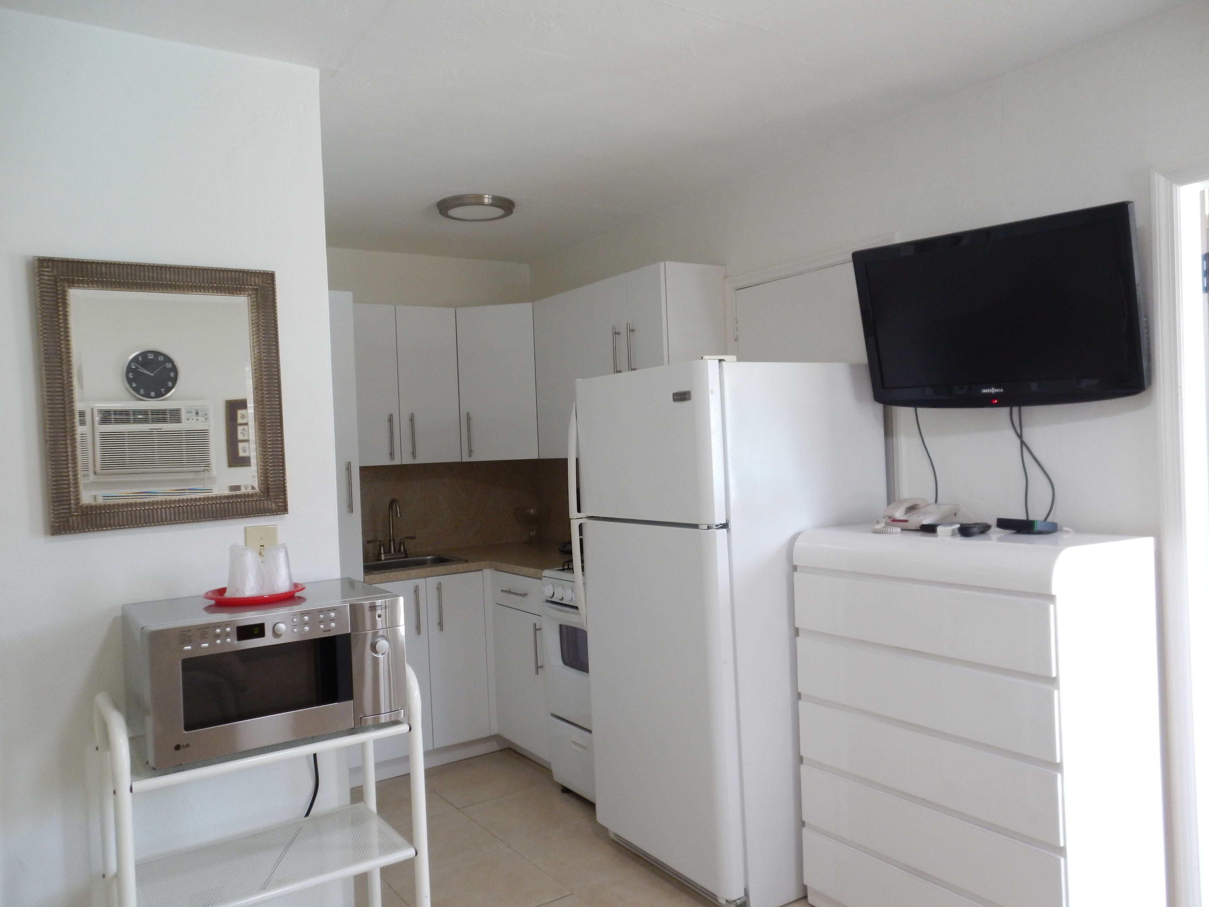 Cocina compacta con armarios blancos, un refrigerador blanco, microondas y un horno pequeño. Un televisor está montado en la pared sobre la encimera.