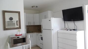 Cocina compacta con armarios blancos, un refrigerador blanco, microondas y un horno pequeño. Un televisor está montado en la pared sobre la encimera.
