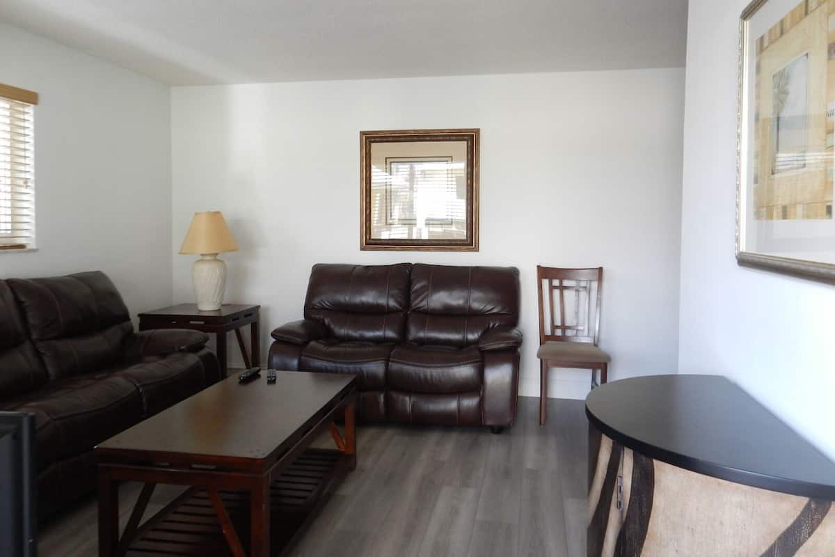 A living room with a brown leather sofa, a wooden coffee table, a small wooden side table with a lamp, and a framed picture on the wall.