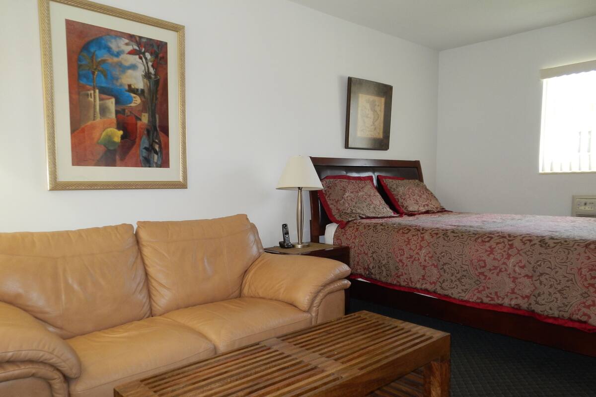 A hotel room with a leather sofa, a wooden coffee table, a bed with patterned bedding, a nightstand with a lamp, and a framed artwork on the wall.