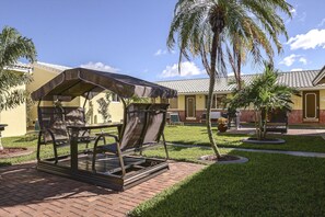 A patio area with a covered seating arrangement, a palm tree, and a building with a tiled roof. - Shell Motel (Hollywood)