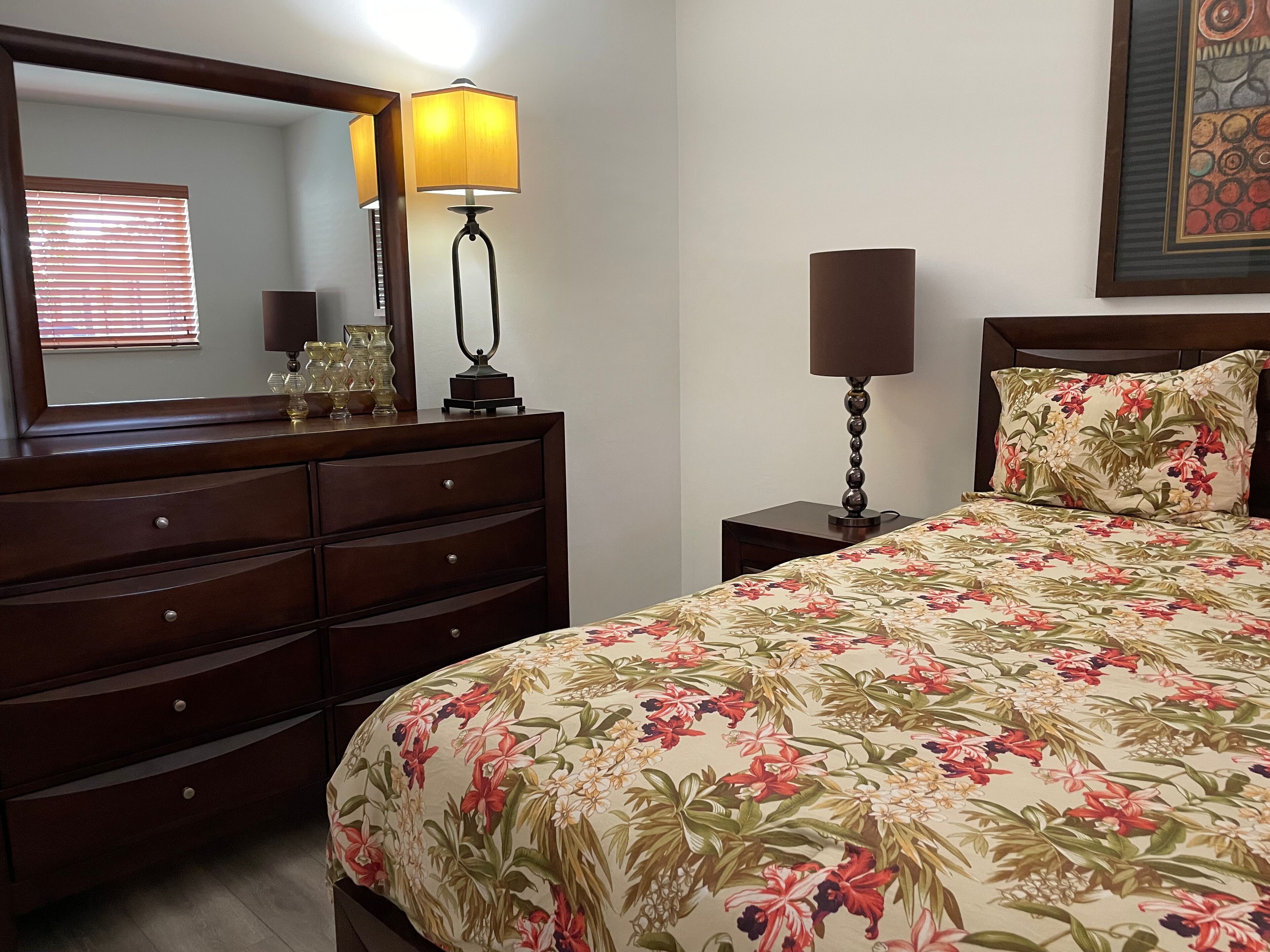 Un dormitorio con un aparador de madera, una cama con ropa de cama floral, una mesita de noche con una lámpara y un espejo montado en la pared.