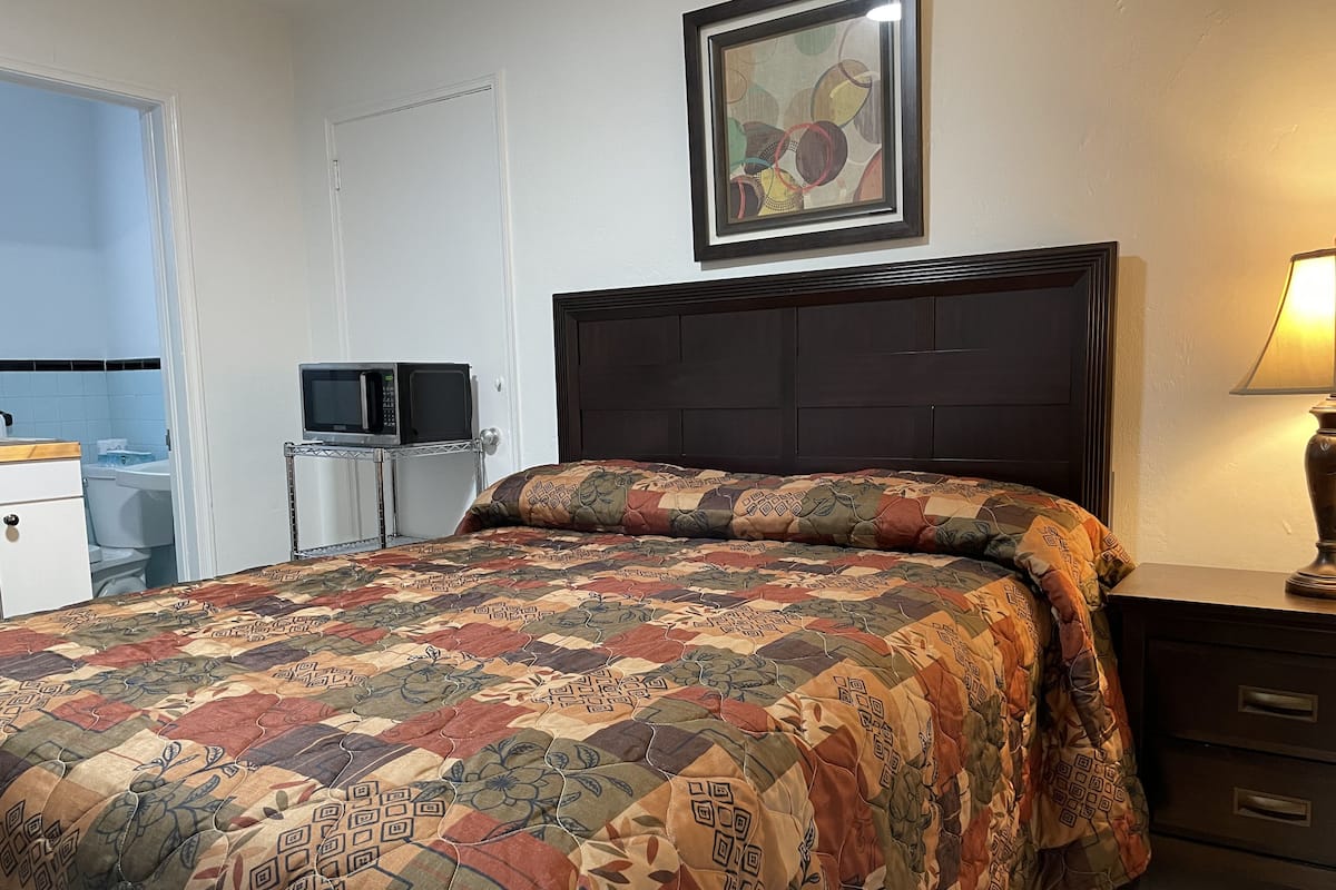 A bedroom with a large bed, a wooden headboard, a bedside table with a lamp, a framed picture on the wall, and a small desk with a microwave.