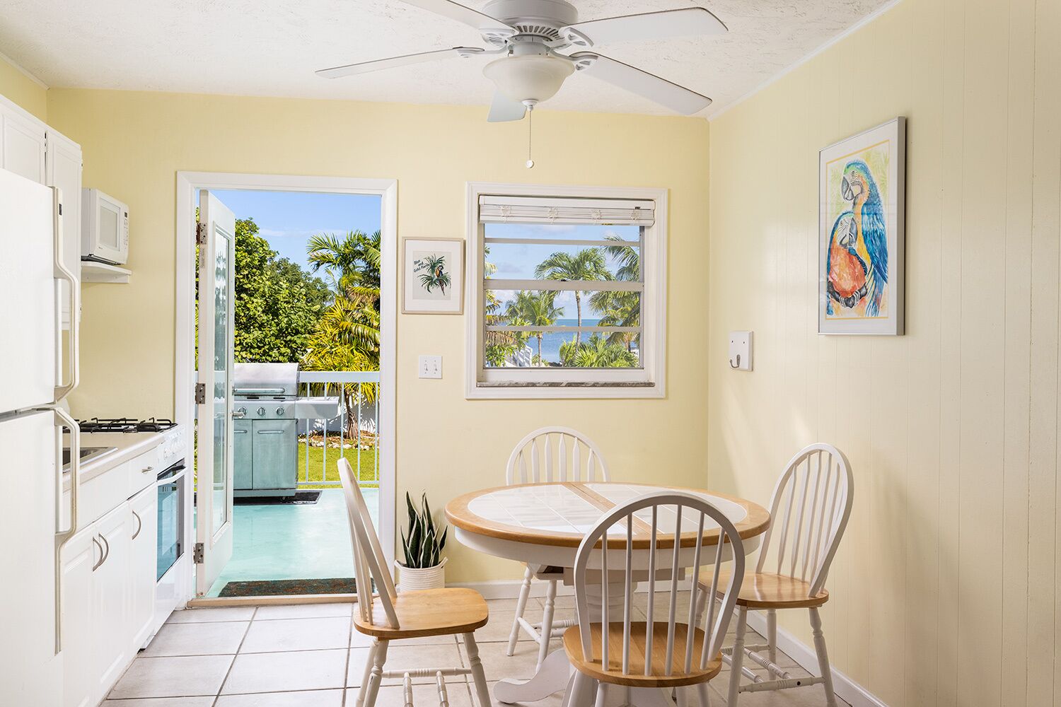 A compact kitchen with white cabinets, a dining area with a round table and four chairs, a ceiling fan, and a window with a view of palm trees and the ocean.