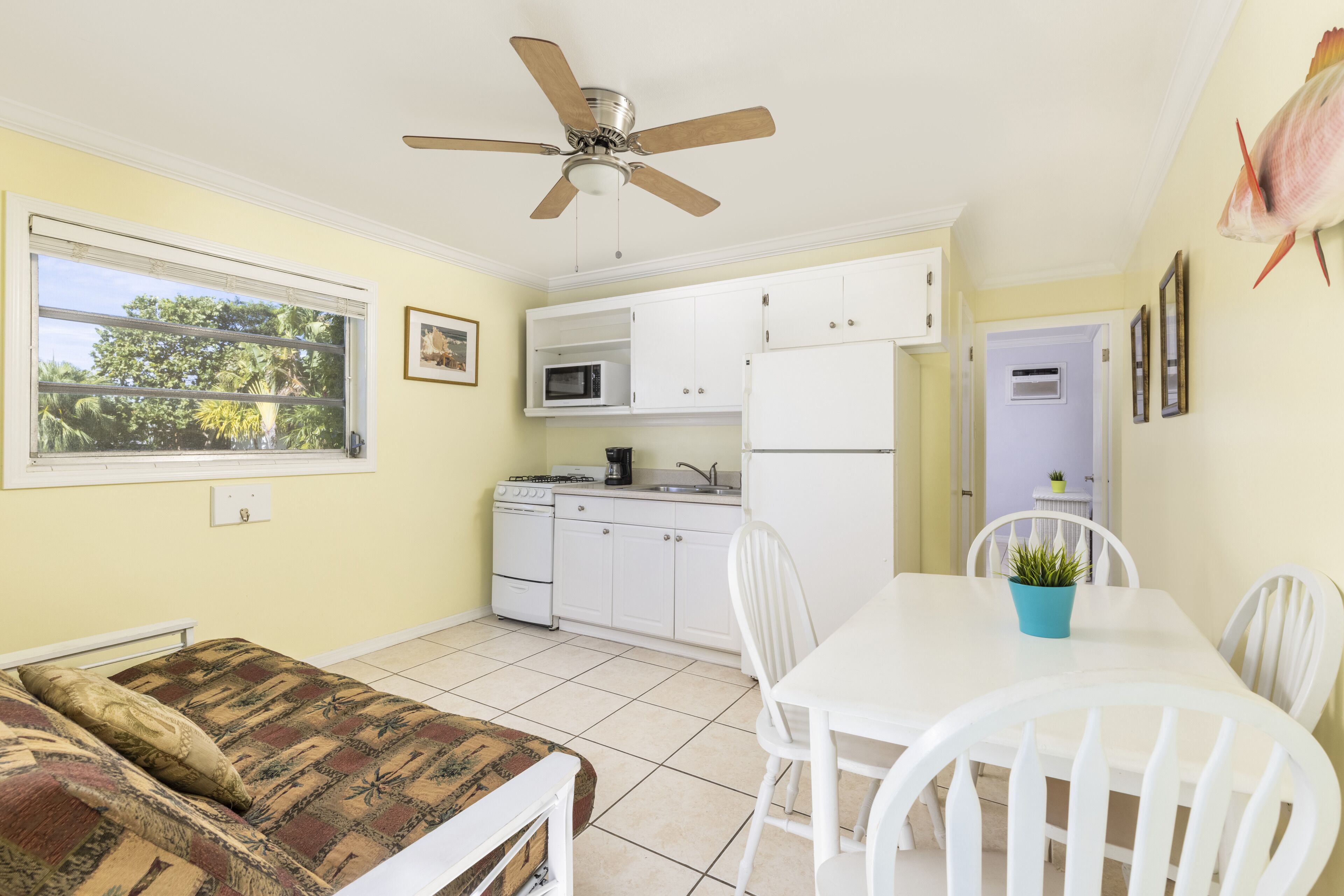 A compact kitchen with white cabinets, a microwave, and a refrigerator. There is a dining table with chairs and a ceiling fan above.